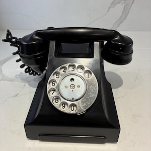 Puede incluir: Un teléfono de disco negro con un dial circular plateado que muestra números negros del cero al nueve. El dispositivo incluye un auricular negro a juego apoyado en la parte superior, conectado por un cable negro en espiral en el lado izquierdo.