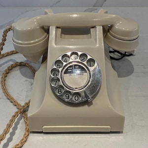 May include: A cream-coloured rotary telephone with a matching handset resting on top. The circular metal dial features black numbers from 1 to 0 and a centre label with text including Fire, Police, Ambulance, and Operator. A braided cord is attached.
