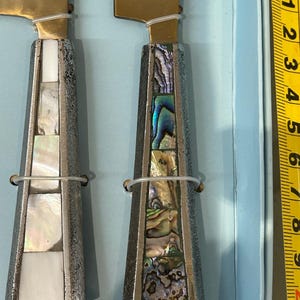 K&ouml;nnte beinhalten: Zwei K&auml;semesser mit goldfarbenen Klingen und dekorativen Griffen. Die Griffe sind silberfarben mit eingelegten Abschnitten aus wei&szlig;em und schillerndem Abalone-Muschel. Ein durchsichtiges Kunststoffband sichert die Klinge am Griff.