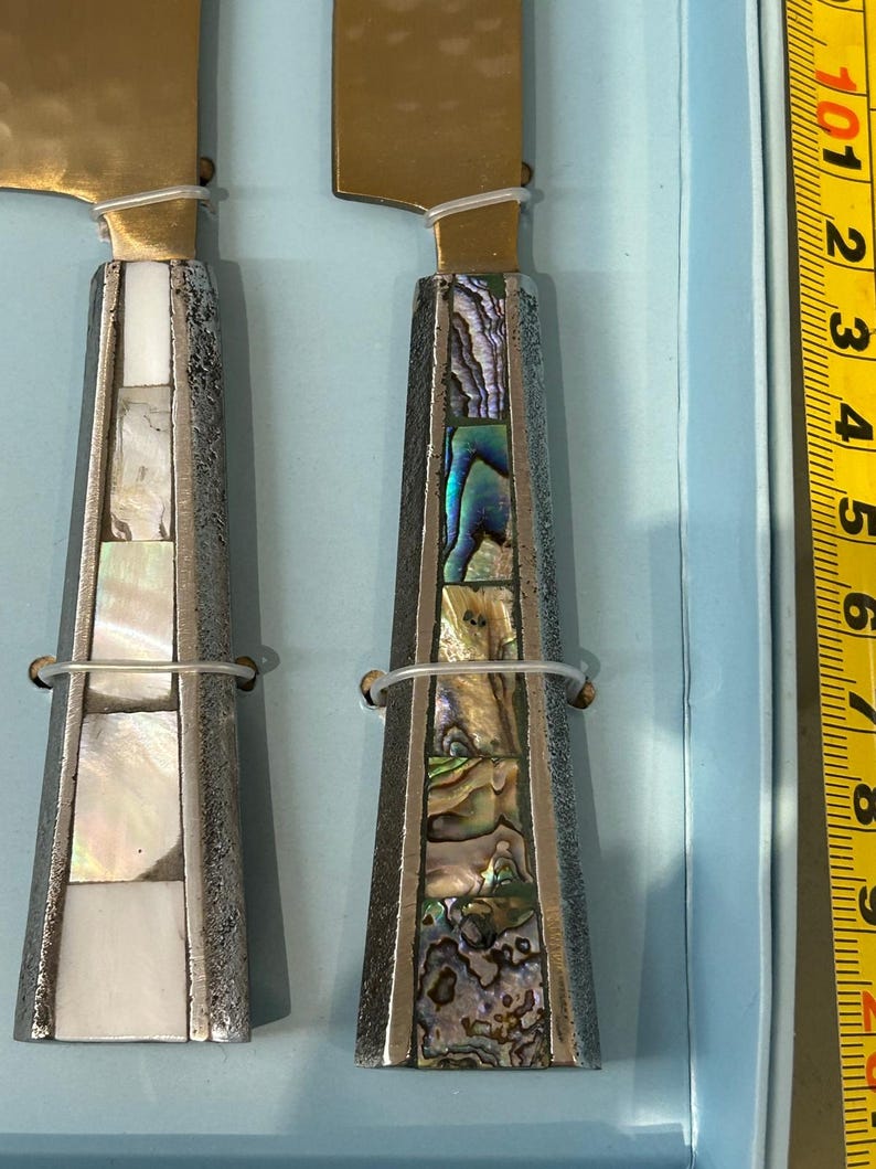 K&ouml;nnte beinhalten: Zwei K&auml;semesser mit goldfarbenen Klingen und dekorativen Griffen. Die Griffe sind silberfarben mit eingelegten Abschnitten aus wei&szlig;em und schillerndem Abalone-Muschel. Ein durchsichtiges Kunststoffband sichert die Klinge am Griff.