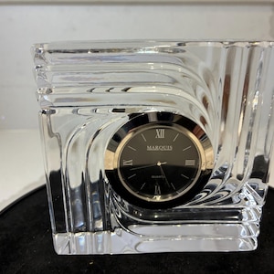 Puede incluir: Un reloj de cristal transparente con esfera negra y ribete dorado. La esfera tiene la palabra "MARQUIS" y números romanos. El reloj tiene forma cuadrada con un diseño decorativo en espiral.