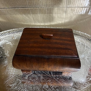 May include: A small, square wooden box with a curved base and a lid with a small handle. The box is a rich, dark brown color and sits on a silver tray with an ornate floral pattern. The box appears to be vintage.