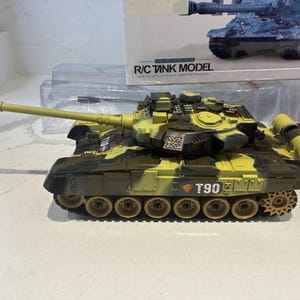 Könnte beinhalten: Ein ferngesteuertes T90-Panzermodell mit Tarnmuster in Olivgrün, Beige und Schwarz. Der Panzer verfügt über ein langes Geschützrohr, einen drehbaren Turm und sichtbare Ketten. Die Verpackung zeigt den Schriftzug R/C TANK MODEL.