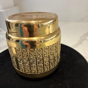 Peut inclure: Un pot doré avec un couvercle et des volutes répétées sur le corps. Le couvercle a un design simple et arrondi. Le pot est posé sur une surface noire. Le pot semble être en métal.