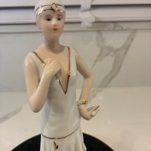 May include: A porcelain figurine of a person with short hair wearing a white dress with gold trim. The figure has a headband, a deep V-neckline, and a ruffled skirt. One hand is placed near the chest, while the other rests on the hip.