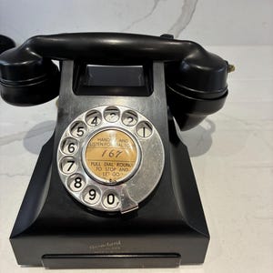 Puede incluir: Un teléfono de disco negro con un dial plateado que muestra los números del cero al nueve. La etiqueta central dice: LIFT HANDSET AND LISTEN FOR DIAL TONE, PULL DIAL ROUND TO STOP AND LET GO, con el número 167 escrito a mano. Standard está impreso.