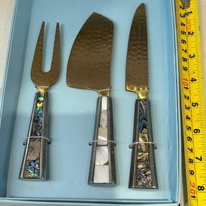 K&ouml;nnte beinhalten: Ein K&auml;semesser-Set mit einer Gabel, einem flachen Messer und einem spitzen Messer. Die Klingen sind goldfarben und die Griffe sind silberfarben mit Abalone- und Perlmutt-Inlays. Das Set befindet sich in einer hellblauen Box. Ca. 20 cm lang.