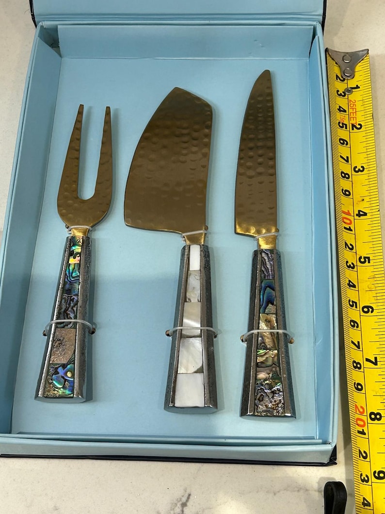 K&ouml;nnte beinhalten: Ein K&auml;semesser-Set mit einer Gabel, einem flachen Messer und einem spitzen Messer. Die Klingen sind goldfarben und die Griffe sind silberfarben mit Abalone- und Perlmutt-Inlays. Das Set befindet sich in einer hellblauen Box. Ca. 20 cm lang.