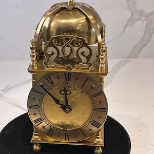 May include: A brass lantern clock featuring a circular face with black Roman numerals and ornate hands. The clock is topped with a dome-shaped bell and a decorative finial, supported by four small ball feet, with intricate metalwork above the dial.