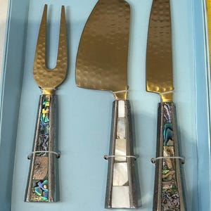 K&ouml;nnte beinhalten: Ein K&auml;semesser-Set mit einer Gabel, einem K&auml;semesser und einem Messer. Die Klingen sind goldfarben mit geh&auml;mmerten Texturen. Die Griffe sind silberfarben mit Abalone- und Perlmutt-Inlays. Das Set wird in einer hellblauen Box pr&auml;sentiert.
