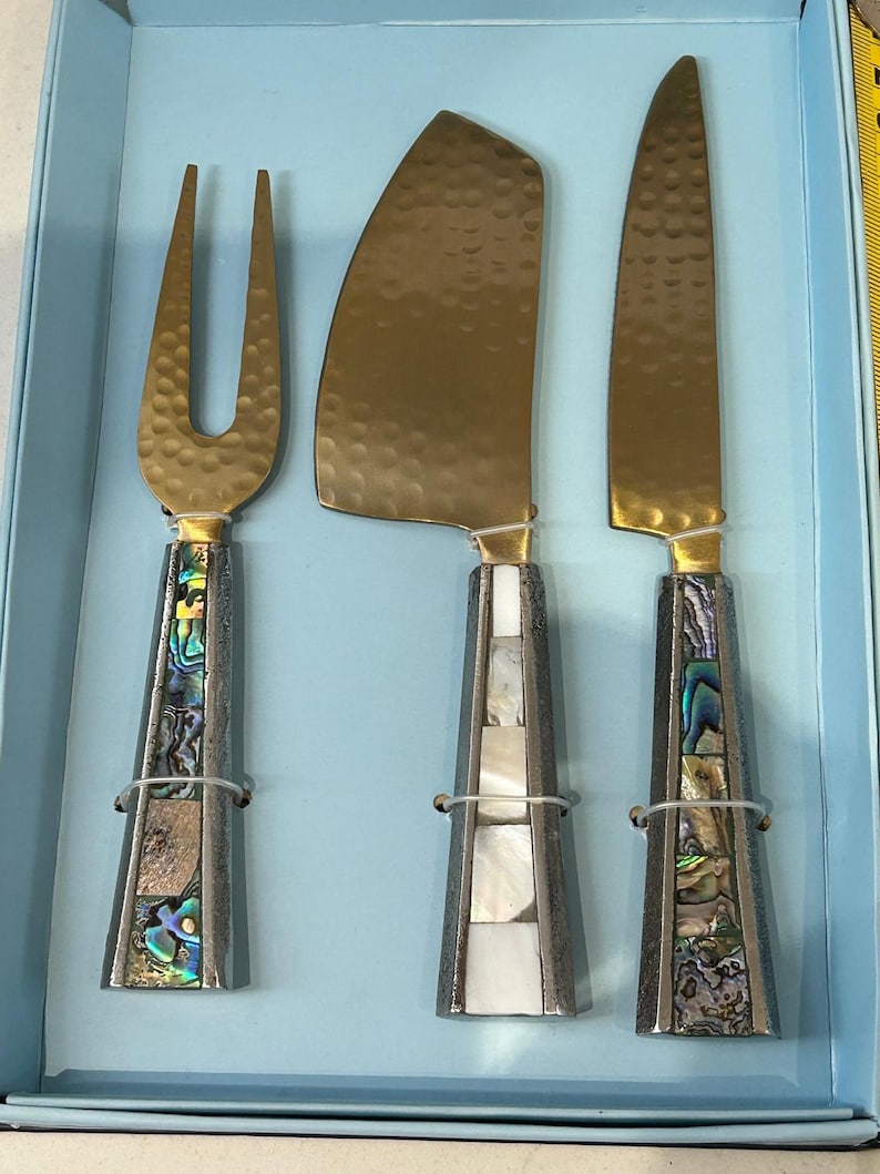 K&ouml;nnte beinhalten: Ein K&auml;semesser-Set mit einer Gabel, einem K&auml;semesser und einem Messer. Die Klingen sind goldfarben mit geh&auml;mmerten Texturen. Die Griffe sind silberfarben mit Abalone- und Perlmutt-Inlays. Das Set wird in einer hellblauen Box pr&auml;sentiert.