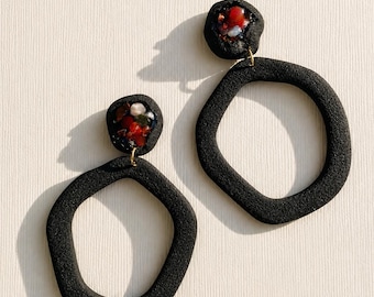 May include: A pair of black earrings featuring a textured, irregular circular hoop suspended from a smaller, rounded stud. The stud contains a cluster of embedded, multi-colored stones in shades of red, white, and dark green with a glossy finish.