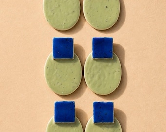 May include: Three pairs of earrings, each featuring a small, glossy, deep blue square component attached to a larger, matte, light sage green oval piece. The surfaces of the oval components have a subtle, textured, speckled appearance.