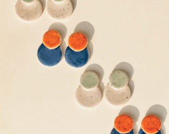 May include: Four pairs of ceramic earrings, each featuring two stacked circles of varying sizes. Two pairs are light beige with speckled textures and pale green tops, while two pairs feature deep blue bases with vibrant orange circular tops.