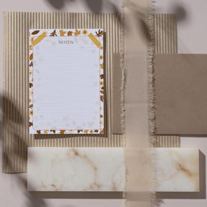 Può includere: Un blocco note a righe con un bordo floreale con foglie e fiori autunnali. La parola "NOTES" è stampata in alto. Il blocco note è posto su uno sfondo strutturato con altri elementi di cancelleria.