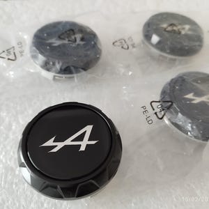 Op de afbeelding: Vier zwarte en zilveren wieldopjes voor auto's, elk in een doorzichtige plastic zak. De doppen hebben een zwarte achtergrond met een wit "A"-logo. De doppen zijn rond met een geribbelde rand.