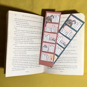 Puede incluir: Dos marcapáginas con ilustraciones de estanterías, plantas y un gato. Los marcapáginas están en un esquema de color rosa y turquesa. Los marcapáginas están colocados encima de un libro abierto.