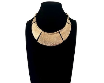 May include: A gold-toned statement necklace with a wide, geometric design. The necklace is made of multiple pieces of metal that are connected together to form a large, rounded shape.