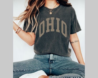 Könnte beinhalten: Ein dunkelgraues T-Shirt mit dem Wort "OHIO" in großer, fetter Schrift gedruckt. Das Shirt wird von einer Person mit langen braunen Haaren getragen und zu einer blauen Jeans kombiniert.