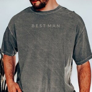Könnte beinhalten: Graues kurzärmeliges T-Shirt mit dem Text "BEST MAN" auf der Brust. Das Shirt hat einen Vintage-Look. Die Person auf dem Bild trägt eine Jeans und eine braune Uhr.