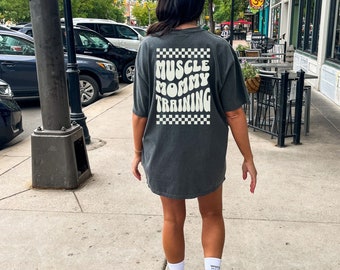 Könnte beinhalten: Ein graues T-Shirt mit einem weißen karierten Rand und dem Text "Muscle Mommy Training" in einer Retro-Schrift.