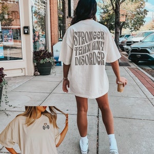 May include: A white oversized t-shirt with the text "Stronger than any disorder" printed on the back. The shirt is being worn by a person walking down a sidewalk. The person is wearing white sneakers.