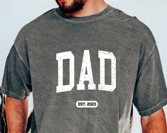 Puede incluir: Una camiseta gris con la palabra "DAD" en letras blancas y el texto "EST. 2023" debajo.