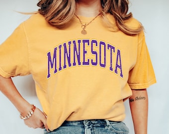 Op de afbeelding: Een vrouw draagt een geel T-shirt met het woord "MINNESOTA" erop gedrukt in paarse en gouden letters. Ze draagt ook een blauwe jeans.