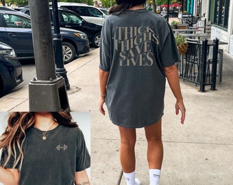 May include: Two women wearing dark gray t-shirts with different designs. The shirt on the left has a dumbbell graphic. The shirt on the right has the text "THICK THIGHS SAVE LIVES".