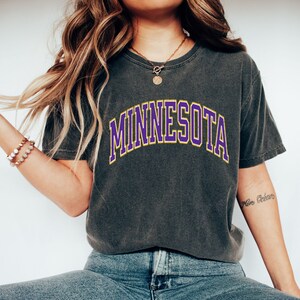 Peut inclure: Un t-shirt gris anthracite avec un graphisme "MINNESOTA" en violet et jaune.