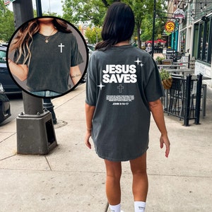 May include: A dark gray oversized t-shirt with the text "JESUS SAVES" and a cross printed on the back. The shirt also includes a bible verse, "For God so loved the world that he gave his only begotten Son, that whosoever believeth in him should not perish, but have everlasting life. John 3:16-17."