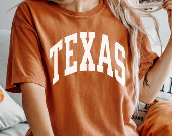 Puede incluir: Una mujer lleva una camiseta naranja quemada con la palabra "TEXAS" impresa en blanco en la parte delantera. Lleva pantalones vaqueros negros rotos y está sentada en una cama.