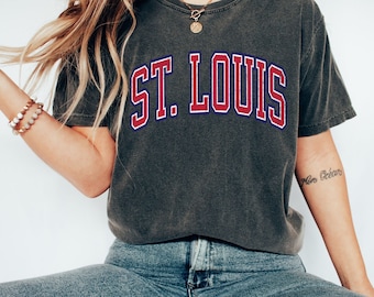 May include: A charcoal gray t-shirt with the text "ST. LOUIS" in red and blue lettering. The shirt is worn by a person wearing blue jeans and white sneakers.