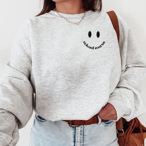 May include: A gray sweatshirt with a black smiley face and the text "school nurse" on the front.
