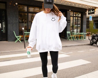 Puede incluir: Una mujer con una sudadera blanca con un gráfico de cara sonriente negra, leggings negros, una gorra de béisbol negra con texto blanco y zapatillas blancas. Lleva una taza de café blanca con un logotipo verde.