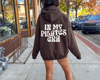 May include: A brown hooded sweatshirt with the text "In My Pilates Era" printed in white on the back.
