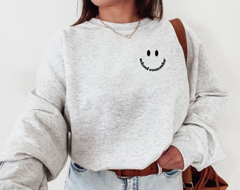 May include: A gray sweatshirt with a black smiley face and the text "school counselor" printed on the front. The sweatshirt is being worn by a person with brown hair and a brown leather bag.