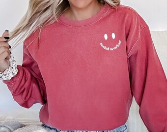 Puede incluir: Una sudadera roja con una carita sonriente blanca y el texto "social worker" debajo.