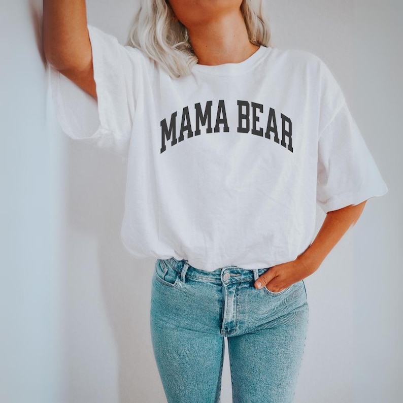 May include: White t-shirt with the text "MAMA BEAR" in black, arched lettering. The shirt is paired with light blue jeans. The image shows a person wearing the shirt and jeans.