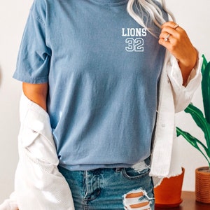 May include: A blue t-shirt with white text that says "LIONS 32" on the front. The shirt is worn by a person wearing denim shorts and a white jacket.