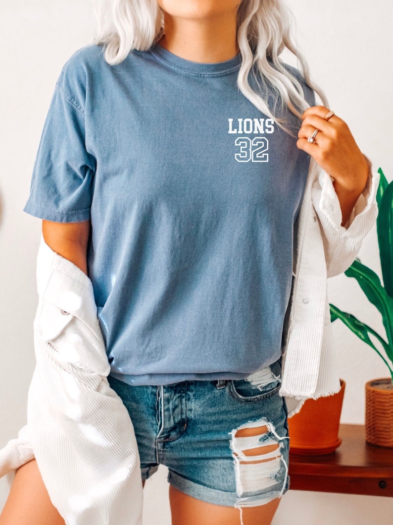 May include: A blue t-shirt with white text that says "LIONS 32" on the front. The shirt is worn by a person wearing denim shorts and a white jacket.