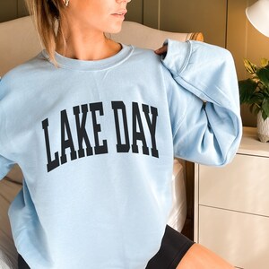 K&ouml;nnte beinhalten: Ein hellblauer Pullover mit dem Aufdruck "LAKE DAY" in Schwarz.