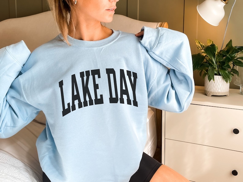 K&ouml;nnte beinhalten: Ein hellblauer Pullover mit dem Aufdruck "LAKE DAY" in Schwarz.