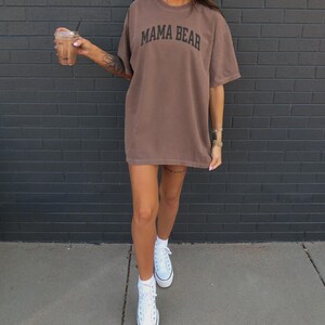 May include: Brown oversized t-shirt with the text "MAMA BEAR" in black. The shirt is paired with white sneakers. The person is holding a beverage in a clear cup with a straw.