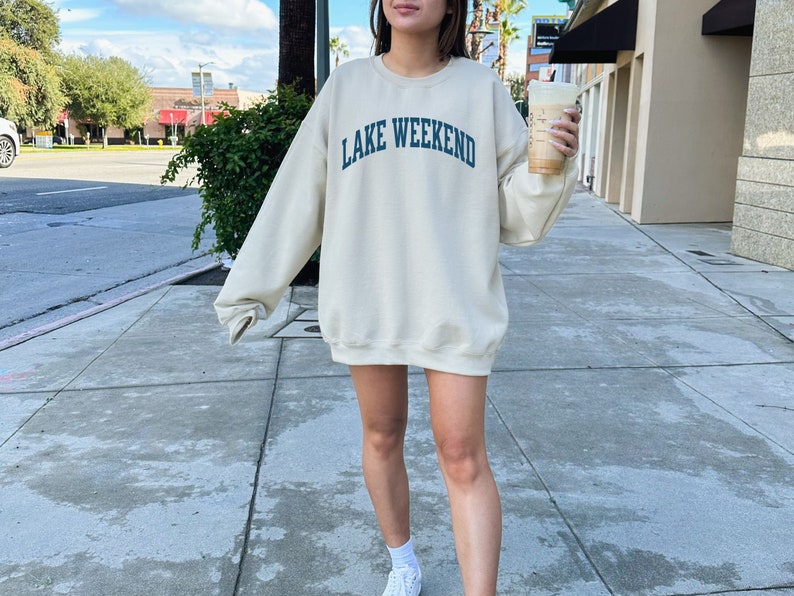May include: A woman wearing a cream colored sweatshirt with the text "LAKE WEEKEND" in blue lettering. She is holding a cup of coffee in her right hand and is standing on a sidewalk.