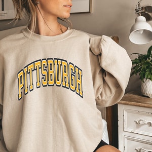 Może przedstawiać: Beżowa bluza z napisem "PITTSBURGH" w ż&oacute;łtych i granatowych literach blokowych. Tekst jest wygięty nad klatką piersiową. Bluza ma okrągły dekolt i długie rękawy. Odzież wykonana jest z miękkiego materiału.