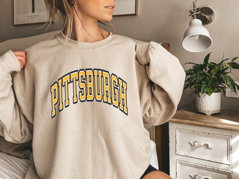 Może przedstawiać: Beżowa bluza z napisem "PITTSBURGH" w ż&oacute;łtych i granatowych literach blokowych. Tekst jest wygięty nad klatką piersiową. Bluza ma okrągły dekolt i długie rękawy. Odzież wykonana jest z miękkiego materiału.