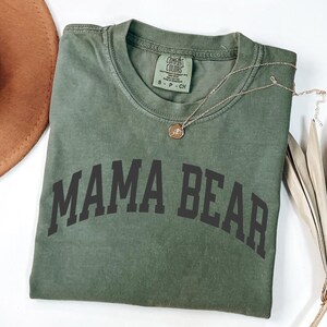 May include: Olive green t-shirt with the words "MAMA BEAR" printed in dark gray. The shirt has a crew neck and short sleeves. A gold necklace with a pendant is draped across the shirt. The shirt is made of a soft, comfortable material.