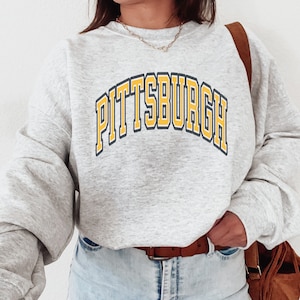 Może przedstawiać: Jasnoszara bluza z napisem "PITTSBURGH" w ż&oacute;łtych i granatowych literach na piersi. Bluza ma długie rękawy i okrągły dekolt. Osoba ma na sobie brązowy pasek i brązową torbę.