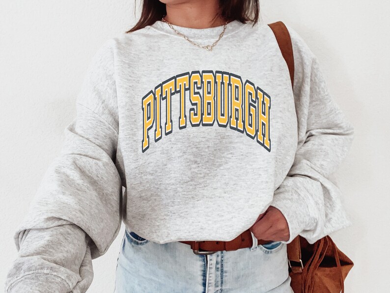 Może przedstawiać: Jasnoszara bluza z napisem "PITTSBURGH" w ż&oacute;łtych i granatowych literach na piersi. Bluza ma długie rękawy i okrągły dekolt. Osoba ma na sobie brązowy pasek i brązową torbę.